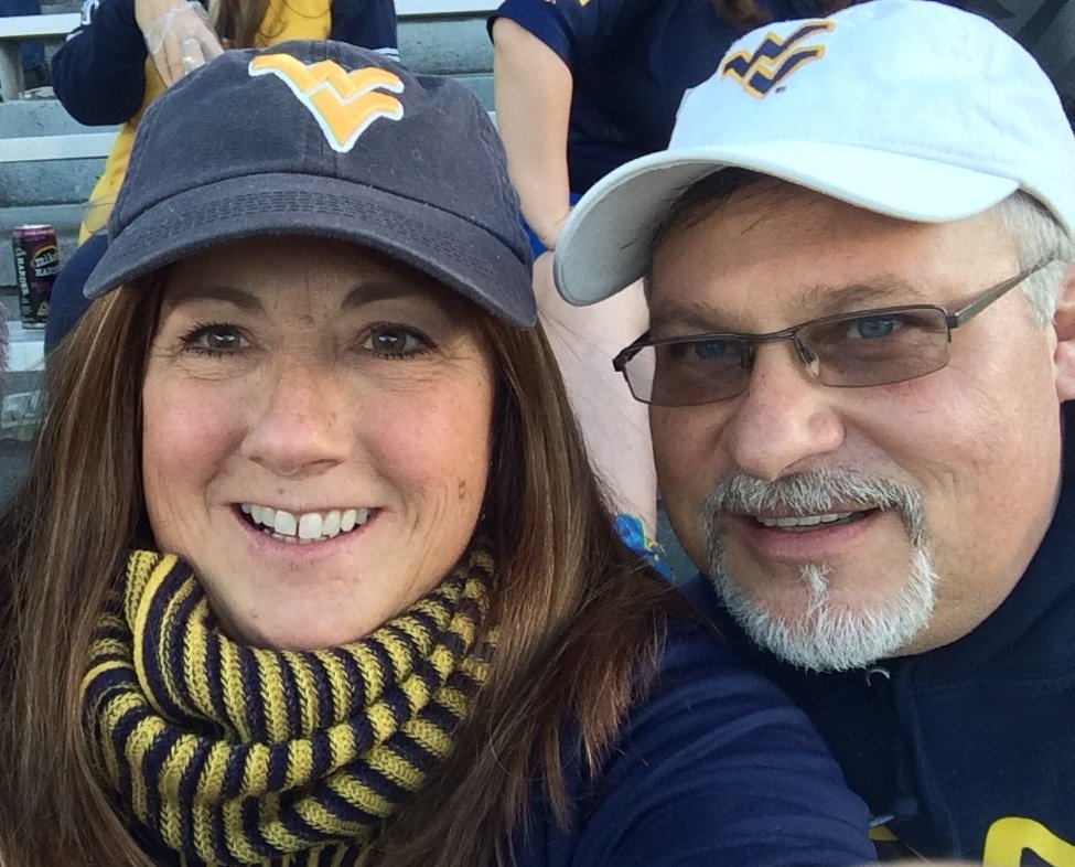 A close-up selfie of a woman and man sitting in stadium bleachers wearing West Virginia University (WVU) gear. The smiling woman on the left wears a navy blue WVU baseball cap and a striped yellow-and-navy scarf. The man on the right wears a white WVU baseball cap, glasses, and has a trimmed goatee.