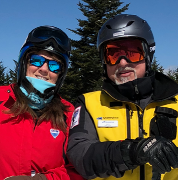 A man and woman smiling together in full ski gear outdoors against a bright blue sky and evergreen trees. The woman on the left wears a red jacket, blue neck gaiter, helmet, and blue reflective sunglasses. The man on the right wears a yellow Snowshoe resort ski jacket, black helmet, and red reflective goggles.
